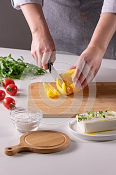 The girl`s hand cuts pieces of pepper on a wooden board, cheese, and tomatoes on a white kitchen table. Preparation of