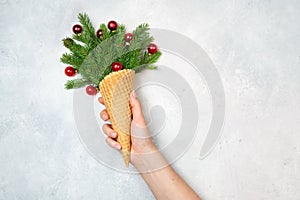 The girl`s hand holds an ice cream cone with sprigs of fir and red Christmas toys. Light gray background. New Year. Christmas