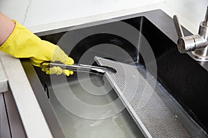 A girl's hand in a yellow glove turns over a filter from a kitchen hood with tongs in the cleaning mixture of an