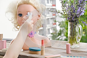 girl's hands are applying makeup on doll, playing with set of stylist, beautician, hairdresser on wooden table in garden, on