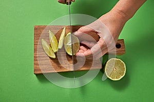 A girl`s hands cut a green natural lime to slices with sharp knife on a wooden board on a green. Top view.