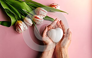 The girl\'s hands hold a white heart on a pink background, as well as tulips and a pink decoration. Holiday concept