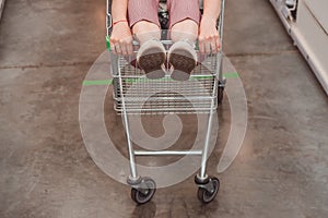 Girl`s legs stick out of the basket in the shopping hall of the supermarket. A teenager rides in a cart in an empty store