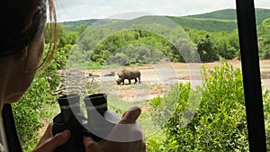 a girl in safari clothes looks at the rhinos through binoculars in the savannah