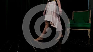 A girl in a salmon-colored dress walks under a dark room with a green chair.