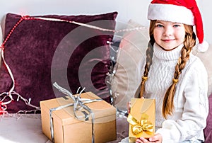 A girl in a santa claus hat sits on the sofa with a new year gift in her hands and looks at the camera