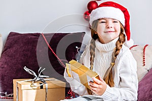 A girl in a santa claus hat sits on the sofa with a new year gift in her hands and looks out the window