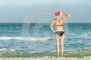 Girl in Santa hat and bathing suit looks into the distance. Inscription New Year on the back. Sea shore.