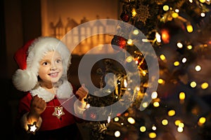 A girl in a Santa hat decorates the Christmas tree on Christmas eve and smiles sweetly in anticipation of the holiday