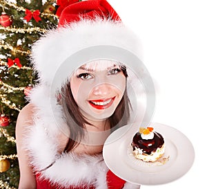 Girl in santa hat eat cake by christmas tree.