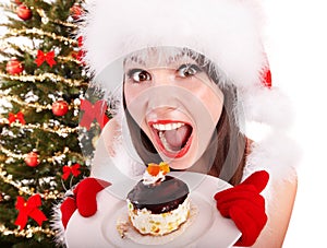 Girl in Santa hat eat cake by Christmas tree.