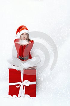 A girl in a Santa hat, sitting on a big red gift box and blowing snow off her palms