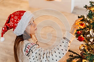 Girl in Santa hat smiling and hanging multi-coloured balls on Christmas tree