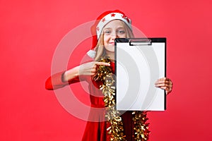 Teen girl in Santa hat and tinsel on neck, holding tablet with clean sheet and pointing finger at him, plans for New year on red