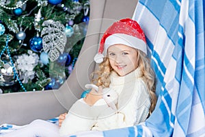 A girl in a Santa hat with a white rabbit at home near the Christmas tree, the concept of new year and Christmas