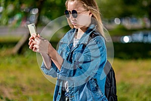 Girl schoolgirl in summer in park. In his hands holds a smartphone. In a denim jacket behind a backpack, sunglasses