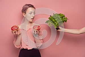 A girl with short hair in a blouse holds donuts in her hands, and looks at the salad in outstretched hand, on a pink