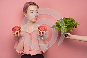 A girl with short hair, holds donuts in her hands and raising an eyebrow, looks at her hand with a salad, on a pink