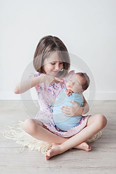 A girl with a short haircut in a pink dress holds a newborn in a blue cocoon, children in a white room