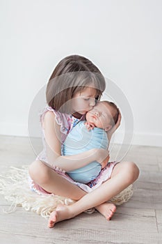 A girl with a short haircut in a pink dress holds a newborn in a blue cocoon, children in a white room