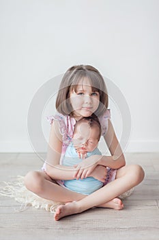 A girl with a short haircut in a pink dress holds a newborn in a blue cocoon, children in a white room