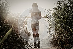 The girl in shorts with rubber shoes standing in lake water with reed nearby and working with tablet PC. Full body view