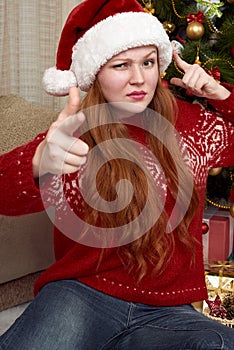 Girl show gesture of a gun. Dressed in retro red sweater and santa hat. Home interior with christmas decoration, fir tree and gift