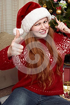Girl show gesture of a gun. Dressed in retro red sweater and santa hat. Home interior with christmas decoration, fir tree and gift