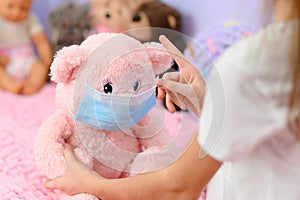 The girl shows her hand to the teddy bear about the importance of wearing a medical mask during coronavirus.