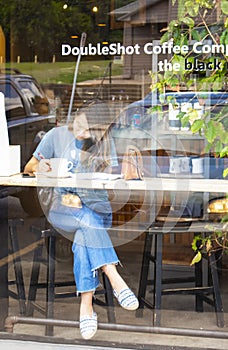 View of girl in coffee shop window working on paperwork with phone laying nearby shot through reflections of outside on glass Doub