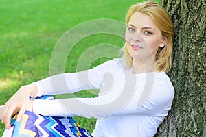 Girl sit on grass lean on tree trunk relaxing in shadow green nature background. Woman blonde take break relaxing in