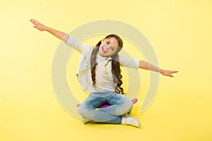 Girl sit penny board yellow background. Child smiling face pretend fly on skateboard. Kid relax sit penny board