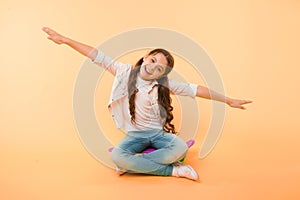 Girl sit penny board yellow background. Child smiling face pretend fly on skateboard. Kid relax sit penny board