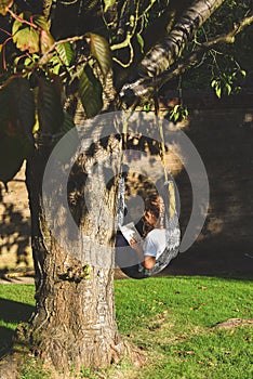 A girl sits alone in a comfy hammock reading a book during a warm sunny evening garden