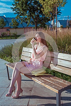A girl sits on a bench in city park in summer, drinks tea from cup of coffee, handbag. Rest after work, lunch at lunch