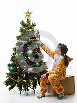 The girl sits on a box in front of the Christmas tree and hangs a Christmas tree ball