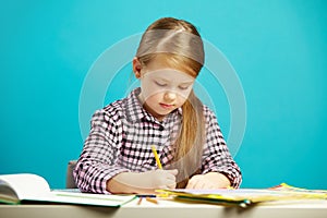 Girl sits at desk and writes to notebook, on blue isolated background. First grader in school.