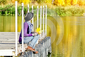 Girl sits on a dock near the water in a hat and with leaves in her hands. Sunny day