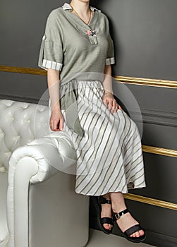 The girl sits on the edge of a white leather sofa in a skirt, blouse and black sandals