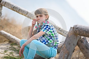 A girl sits on a fence made of a wooden blockhouse and, with a blade of grass in her mouth, looks into the frame