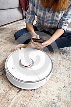 The girl sits on the floor and controls the robot vacuum cleaner using a smartphone, smart home