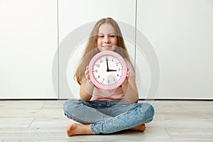 girl sits on the floor and holds a watch in her hands that shows 3 o& x27;clock p.m.