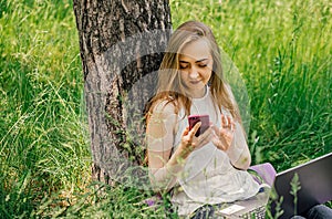 girl sits on the grass and works at a laptop. looks at the phone. freelance. selfeducation. the concept of remote photo