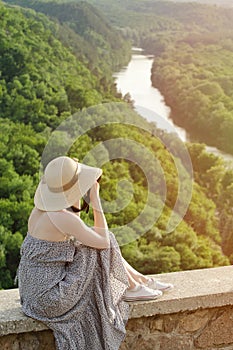 Girl sits on hill and takes pictures against the background of a forest and meandering river