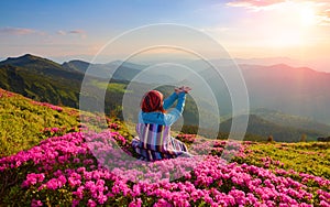 The girl sits on the lawn covered with pink flowers watching at the high mountains, sky with clouds and sun in summer day.