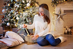 The girl sits near the Christmas tree and opens the golden gifts