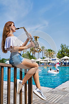 Girl sits near the pool and plays the saxophone on a summer day