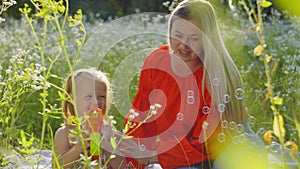 The girl sits next to her mother among the daisies and blows soap bubbles.