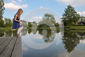 A girl sits on a pole of a boardwalk at a lake with a fishing r