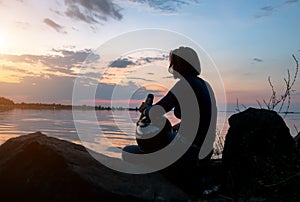 A girl sits on the river bank in the evening. Silhouette of a girl on a sunset background